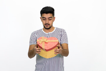 Young man wearing a white striped shirt. He is holding a gift box in the shape of a heart. He has a short beard. White background. Isolated image.