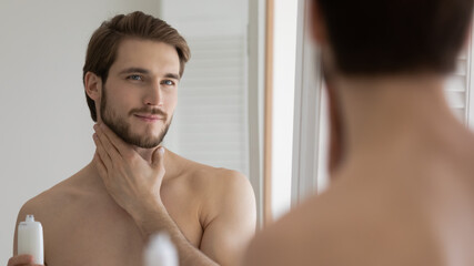 Fototapeta na wymiar Pleasant young handsome caucasian man applying moisturizing lotion, moisturizing facial skin or smoothing after shaving. Well -groomed millennial guy involved in daily skincare routing in bathroom.