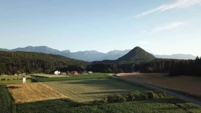 A drone capture of Katherinekogel, towering above the forest in Austria. The small hill resembles a pyramid in shape. In the back there are high Alpine chains. There are many farmlands in front
