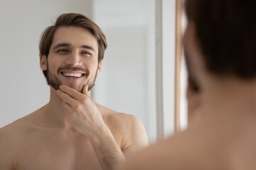 Mirror reflection smiling young 30s handsome well-groomed man standing naked in bathroom, touching...