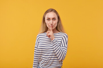 Beautiful young blonde woman looks at the camera and gestures her finger at her mouth. Symbol of silence. Yellow background.