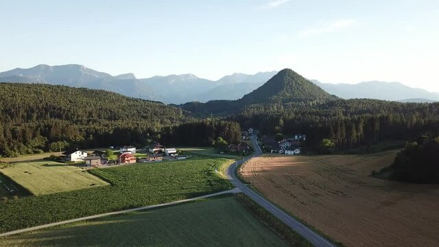 A drone capture of Katherinekogel, towering above the forest in Austria. The small hill resembles a pyramid in shape. In the back there are high Alpine chains. There are many farmlands in front
