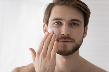 Close up head shot happy young european appearance man looking at camera, applying nourishing or...
