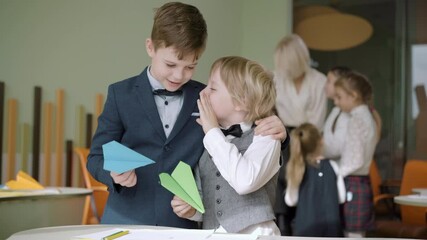 Cute Caucasian boy whispering on ear of friend with blurred classmates talking with teacher at the background. Schoolboys sharing secrets in classroom in public school. Friendship and education. - Powered by Adobe