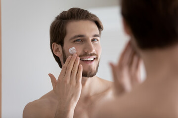 Smiling handsome millennial caucasian man looking in mirror, moisturizing face with nourishing...