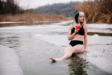 A young sports girl with a cocktail in her hand stands in the winter river in the hole