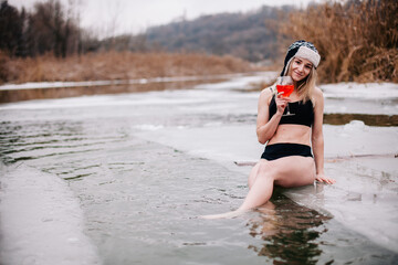 A young sports girl with a cocktail in her hand stands in the winter river in the hole