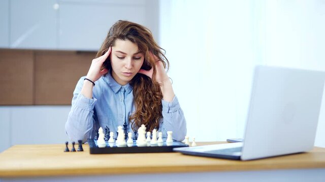 Portrait young attractive woman playing chess game online using laptop at home in the kitchen at the table. Woman during quarantine leisure and hobby play over the internet.