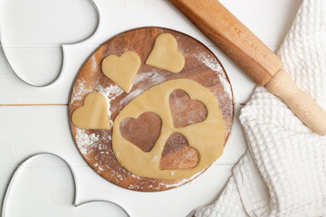 A heart shaped cutout mold and dough cut cookies next to a towel and wooden spoons and rolling pin, flat lay.
