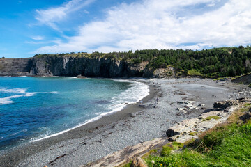 A pebbly beach with a small curve shape. There's a rock face close to the ocean in the distance with trees and jagged coast. Waves are rolling in on the beach. The blue sky is dramatic with clouds.
