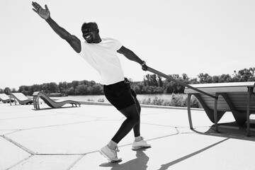 Black man in white t-shirt and black shorts doing exercises using elastic tape on fresh air. Monochrome photo.
