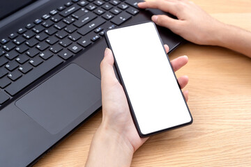 Female hand holding mobile phone with white screen over laptop.