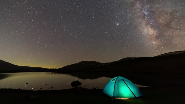 Blue Tent Camping Near the Calm Relax Lake like a Mirror Lantern Light Inside the Camping and Milky Way Nebula Move in Starry Night Sky