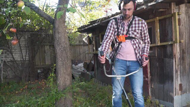 Man with electric lawnmower, lawn mowing. Gardener trimming garden. Suburb, village. Young caucasian male farmer gardener standing in yard with petrol trimmer. Brush cutter cutting weeds on farm