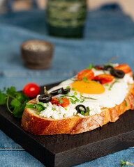 Fototapeta na wymiar Toasted bread toast with fried eggs with yellow yolk and tomatoes, olives, sprinkled with herbs on dark wooden serving board on blue denim napkin, vertical