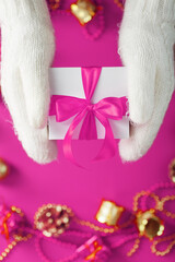 Hands in warm white knitted mittens hold a white gift box with a pink bow on a pink background.