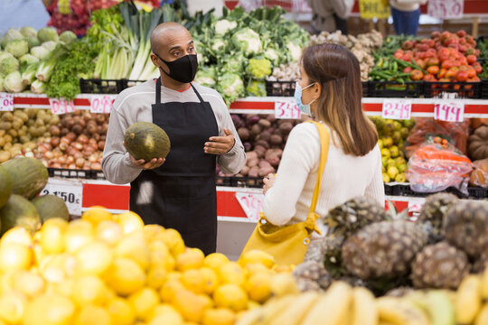 Store Employee Helping Girl Choosing Fruit In Supermarket. People In Medical Masks During COVID-19