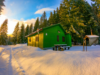 APATIŠAN, VELEBIT MOUNTAIN, CROATIA, December 2020 - Green wooden mountain hut at the edge of the coniferous forest. Beautiful snow day. Wooden table and benches in front of the house 