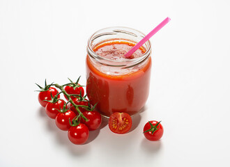 freshly squeezed tomato juice in a glass jar with a cocktail tube, next to there are bright red cherry tomatoes on a branch against the background of a white table.