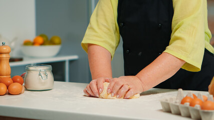 Grandmother hands preparing homemade cookies in modern kitchen kneading on the table. Retired elderly baker with bonete mixing ingredients with wheat flour for baking traditional cake and bread
