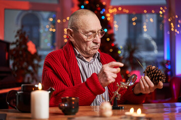 Portrait of a concentrated and serious santa claus working at table and preparing decoration from cone in colourful and decorated cosy room.