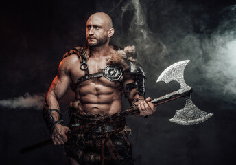 Fototapeta na wymiar Dressed in antique light armour with fur bald and violent viking with muscular build poses in dark atmospheric background