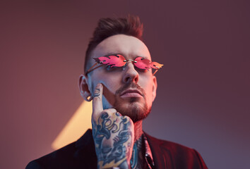 Obraz premium Serious and tattooed hipster businessperson with beard and stylish haircut poses in dark room with lights and colourful walls touching his face with finger.