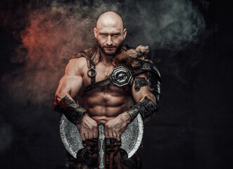 Fototapeta na wymiar Powerful and shirtless viking warrior with bald head dressed in steel lights armour with fur poses in dark atmospheric background