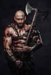Fototapeta na wymiar Serious and furious barbarian with bald head and grimy and bloody skin in armour with fur posing in dark background holding two handed double axe