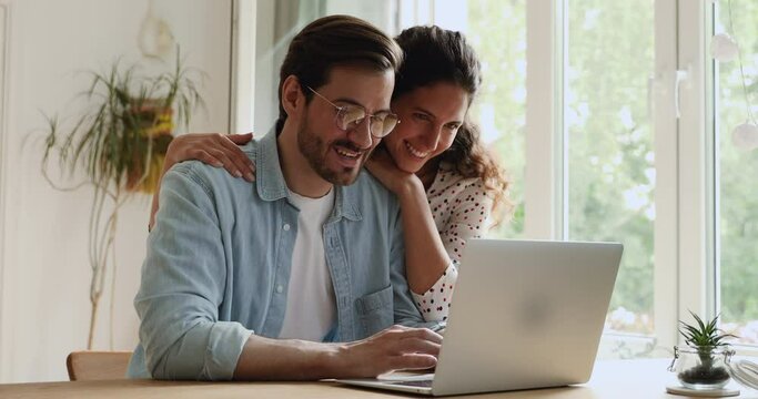 Loving millennial caucasian bonding couple using computer, planning honeymoon or summer vacation, watching family photos, web surfing information or enjoying online shopping together at home.
