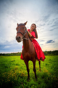 A woman in a bright Gypsy dress and image with a horse in a field with green grass. A model or actress posing in nature with an animal from a farm and the sky with clouds in the background