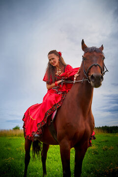 A woman in a bright Gypsy dress and image with a horse in a field with green grass. A model or actress posing in nature with an animal from a farm and the sky with clouds in the background