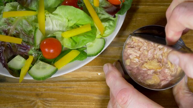 Closeup POV overhead shot of a man&rsquo;s hands removing a can opener then the lid from a tin of chopped skipjack tuna in water, next to a plate of salad, on a pine worktop / counter.