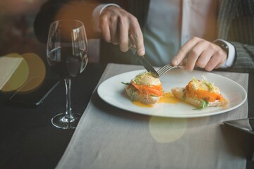 A young and attractive man uses his phone while delicious eggs benedict with lightly salted salmon and sauce in an indoor restaurant