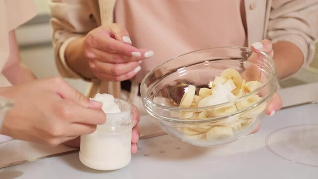 Midsection shot of unrecognizable females adding yoghurt and jam to bowl with cut bananas for homemade ice-cream