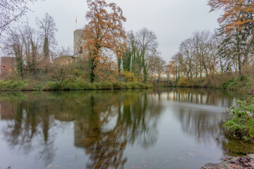 Fototapeta na wymiar Lake surrounded by autumn trees reflected in the water surface, the ruins of Stein Castle in the background, cloudy foggy day in a nature reserve, South Limburg, Netherlands. Long exposure photography
