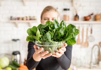 Young blond smiling woman holding a bowl of fresh spinach in the kitchen