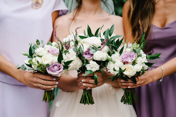 Obraz na płótnie Canvas The bride and bridesmaids in an elegant violet dress is standing and holding hand bouquets of pastel pink flowers and greens at nature. Young beautiful girls holds a wedding bouquet outdoors.