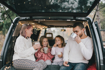Happy parents and their kids drinking hot cocoa sitting in a van decorated with festive Christmas lights.