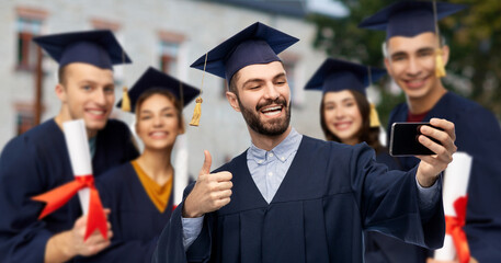 education, graduation and learning concept - happy smiling male graduate student in mortar board and bachelor gown with smartphone taking selfie over group of people on school background