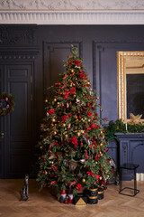 Fototapeta na wymiar a tall, beautifully decorated Christmas tree stands against the background of a dark gray wall and a mirror in a golden frame, a wreath with snow-covered cones hangs on the doors