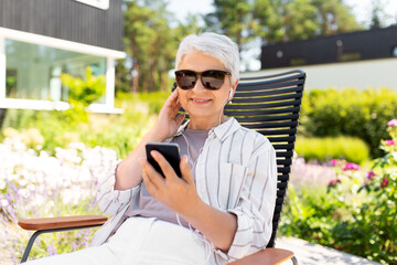 Fototapeta premium old age, retirement and people concept - happy senior woman in sunglasses with earphones and smartphone listening to music at summer garden