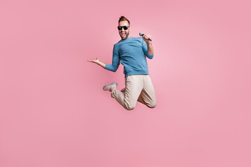 Full length photo of handsome cute guy wear blue pullover black glasses jumping singing holding arm...