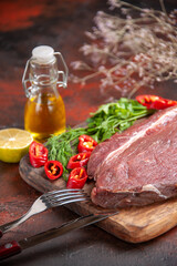 Side view of red meat on wooden cutting board and garlic green chopped pepper fallen oil bottle lemon fork knife on dark background