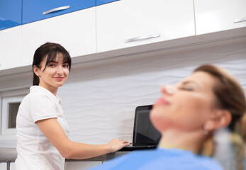 A smiling young dentist at the dentistry office working on laptop and consulting a patient on a dental chair