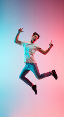 Fototapeta na wymiar Pointing up, shouting. Young caucasian man's jumping on gradient blue-pink studio background in neon light. Concept of youth, human emotions, facial expression, sales, ad. Full length, copyspace.