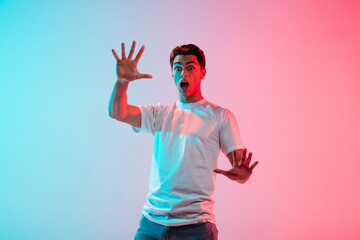 Naklejka premium Scared, shocked. Young caucasian man's portrait on gradient blue-pink studio background in neon light. Concept of youth, human emotions, facial expression, sales, ad. Half length, copyspace.