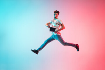 Fototapeta na wymiar Tablet's screen. Young caucasian man's jumping high on gradient blue-pink studio background in neon light. Concept of youth, human emotions, facial expression, sales, ad. Full length, copyspace.