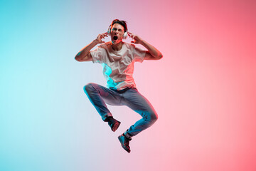 Fototapeta na wymiar Listening to music. Young caucasian man's jumping on gradient blue-pink studio background in neon light. Concept of youth, human emotions, facial expression, sales, ad. Full length, copyspace.