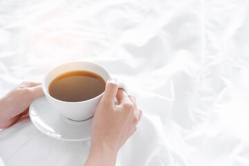 white coffee cup hold by woman hand on bedsheet in weekend with sun light. concept wake up in morning with warm drink to awake self in vacation or holiday relax time in bedroom with copy space.